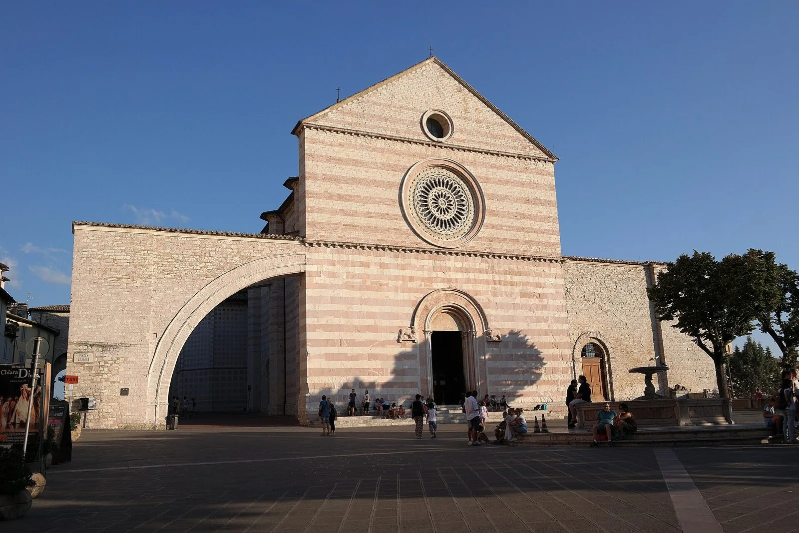 La facciata a fasce di pietra bianca e rosa della Basilica di Santa Chiara ad Assisi, con il grande rosone e l'arco rampante laterale