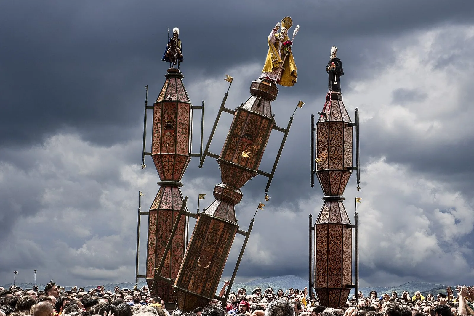 I tre Ceri di Gubbio sormontati dalle statue di San Giorgio, Sant'Ubaldo e Sant'Antonio Abate sollevati sopra la folla, sotto un cielo nuvoloso